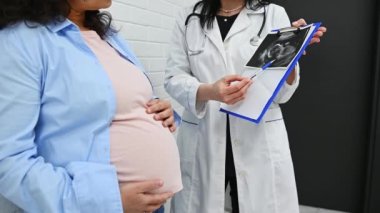 Kadın bir jinekoloğun elinde analiz ve ultrason sonuçları olan bir pano tutarken çekilmiş orta bölüm videosu hamile bir kadına doğum kliniğinde yapılması gereken tedaviyi anlatıyor.