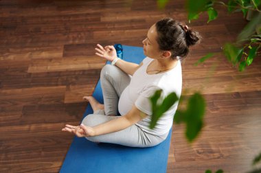 Genel bakış. Latin Amerikalı hamile kadın tozlu ve beyaz tişörtlü, nilüfer kaplamalı yerde oturuyor gözleri kapalı, parmaklarıyla mudra işareti yapıyor, zen, denge ve ahenge uzanıyor.