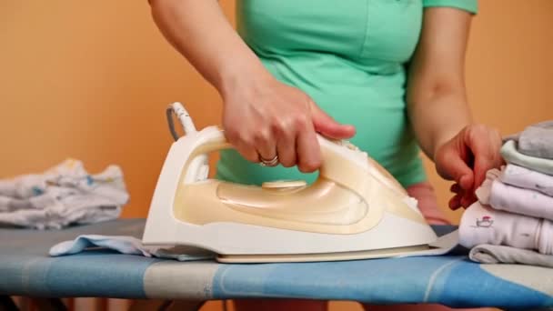 有选择地关注一名孕妇手握电热蒸汽熨斗 熨烫清洗干净的新生儿衣服 将它们折叠在熨烫板上 为妇产医院准备袋子 — 图库视频影像