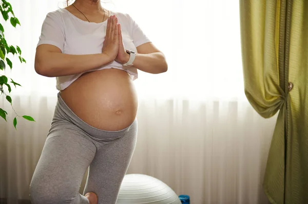 Pregnancy yoga. Expectant gravid female yogi, pregnant woman with naked belly, training tree position with prayer hands at home, practicing meditation and breathing exercises. Copy Space. Cropped view