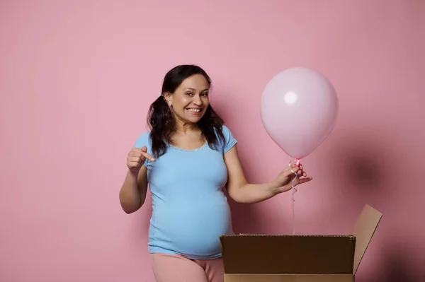 stock image Happy pregnant woman in blue t-shirt, pointing finger at her big belly, holding a pink air balloon. Expectant mom waiting for a baby girl. Gender reveal party. Baby shower. Isolated pink background