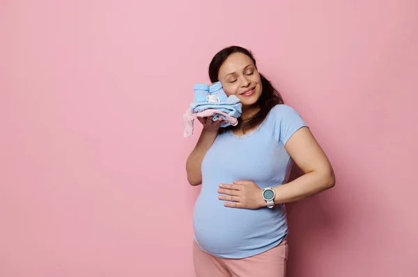 stock image Happy pregnant woman, gently caressing her belly, touches her face to the clothes of future baby, enjoying her pregnancy and maternal lifestyle, expressing positive emotions, isolated pink background