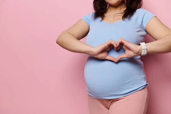 stock image Focus on the hands of a pregnant woman, expectant gravid mother in blue t-shirt, showing heart shape made with her fingers over her big belly, expressing love, tenderness, isolated on pink background