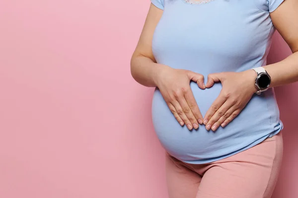 stock image Close-up pregnant woman holding hands on her belly, forming a heart shape with fingers, isolated pink background. Copy space. Pregnancy. Maternity. Childbirth. Gynecology and womens health concept