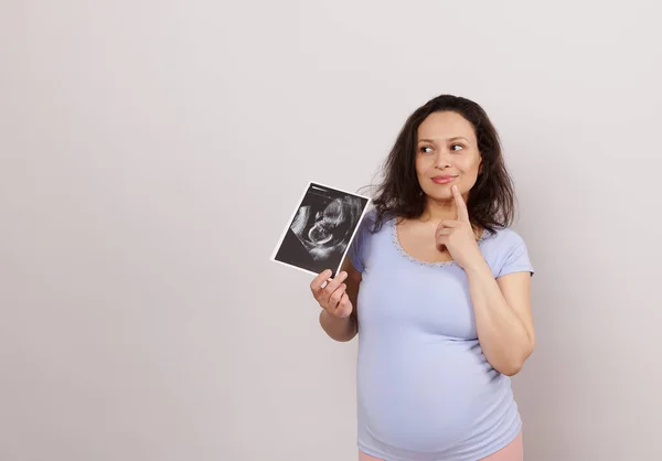 Emotional Latin American pensive pregnant woman, holding ultrasound scan of her baby, dreamily looking aside a copy space on white background. Pregnancy diagnostics concept. Obstetrics and gynecology