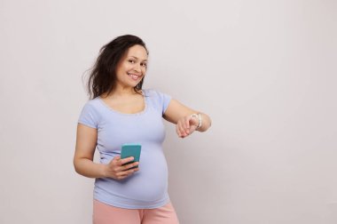 Hamileliği 28 hafta süren mutlu orta yaşlı hamile kadın, bebek bekliyor, akıllı telefon tutuyor ve elektronik akıllı kol saatindeki mobil uygulamaları kontrol ediyor, kameraya bakıp gülümsüyor, beyaz arka planda.