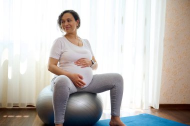 Güzel kıvırcık saçlı, hamile bir kadın karnını tutuyor, bir fitness topuna oturuyor, gülümsüyor, yan bakıyor, doğum öncesi kondisyonunun tadını çıkarıyor. Hamilelikte aktif sağlıklı yaşam tarzı kavramı