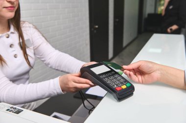 Modern tıp kliniğinin resepsiyon masasında çalışan hoş bir bayan yönetici tarafından tutulan okuyucuya kredi kartıyla ödeme yapan ve şifreli giriş yapan müşterilerin yakın çekim görüntüsü. NFC teknolojisi.