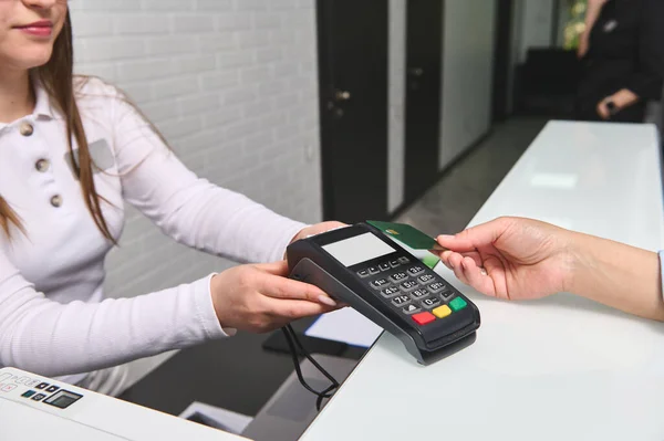 stock image Close-up view of customers hand paying by credit card and entering pin code on reader holded by a pleasant female manager administrator in the reception desk of modern medical clinic. NFC technology.