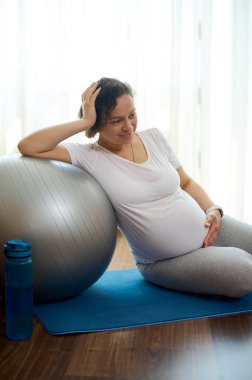Gravid kadın hamile karnının üzerinde elini tutuyor, doğum öncesi yoga ve meditasyondan sonra egzersiz minderinde dinleniyor, hamilelik zamanında sağlıklı ve aktif bir yaşam tarzının olumlu duygularını ifade ediyor.