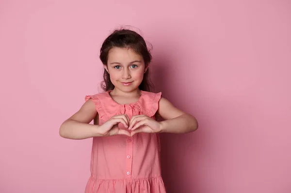 stock image Adorable beautiful Caucasian blue eyed little child girl in pink dress, forms a heart shape with her hands, smiling looking at camera, expressing positive emotions, isolated pink background Copy space