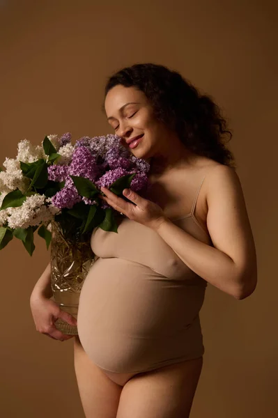 stock image Delightful gravid woman mother in beige lingerie, posing with purple and while blooming lilacs, isolated on beige background. Pregnancy. Maternity. Gynecology. Womens health and fertility concept