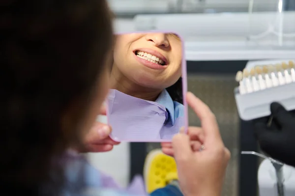 Beautiful smile of female patient, looking at her mirror reflection, after teeth bleaching procedure in dentistry clinic. Cropped view of dentists hands hold a dental scale with teeth color and shade
