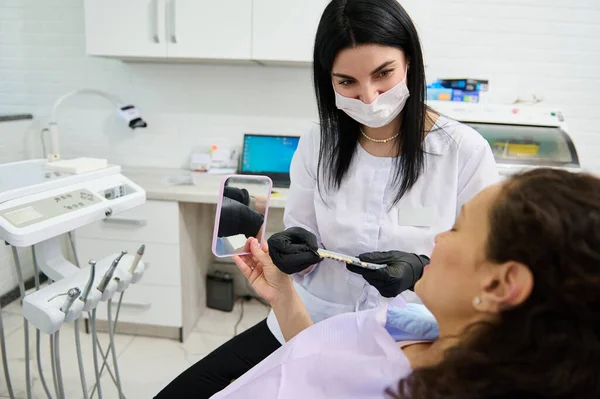 Smiling beautiful female dentist in medical mask holds teeth color chart according to Vita scale, discussing with patient in dental chair the teeth prosthesis and bleaching in modern dentistry clinic