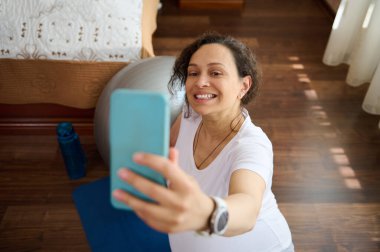 Güzel hamile bir kadın, mavi egzersiz paspasına oturmuş gülümsüyor, akıllı cep telefonuyla selfie çekiyor, hamileliğinin ve sosyal medyada annelik hayatının mutlu anlarını paylaşıyor.