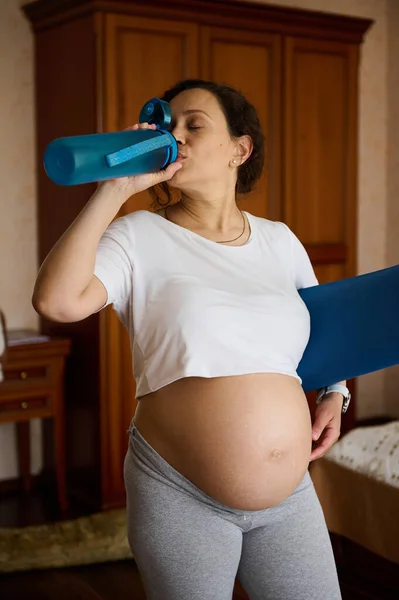 妊娠中のスポーティな女性は水を飲み フィットネスワークアウト後のアクアバランスを更新し 運動マットを保持し 自宅のインテリアに立って 妊娠や産休で活発な健康的なライフスタイルを楽しむ — ストック写真