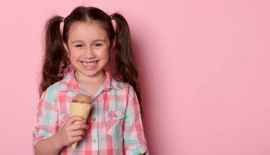 5-6 yaşlarında, şık ekose gömlekli, iki at kuyruklu, gülümseyen, kameraya bakan, waffle külahında dondurma tutan, izole edilmiş pembe arka planı olan yaramaz beyaz bir kız çocuğu.