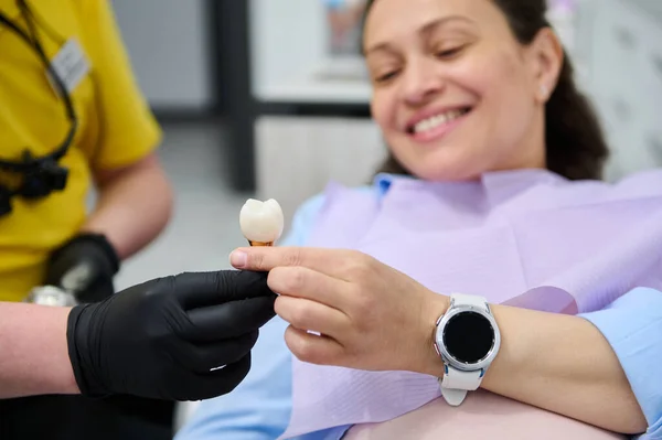 Selective focus on veneer dental implant sample, in hands of a dentist doctor orthodontist showing dentures - prosthesis and explaining treatment to patient, during dental check up in dentistry clinic