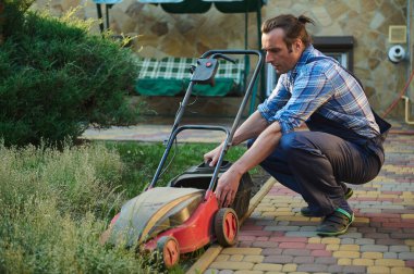 Profesyonel erkek bahçıvan mavi bahçıvan, filtreden kesilmiş yeşil çimenleri çıkarır, evin arka bahçesindeki elektrikli çim biçme makinesini gübrelemek ve şarj etmek için.