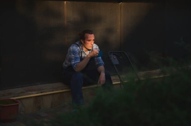 Ön plandaki çitlerin arasından elinde bir fincan kahveyle çalışan genç bir erkek bahçıvana bakın, gün batımında bahçecilikte ve peyzajda sıkı çalıştıktan sonra mola veriyor. Halk ve İşçi Konsepti