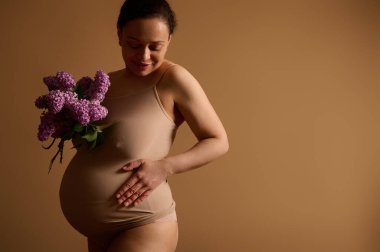 Çekici hamile kadın gülümsüyor, karnına dokunuyor, hamileliğinin mutlu anlarından zevk alıyor, bej iç çamaşırı giymiş, stüdyo arka planında izole edilmiş, mor çiçek açan leylaklarla poz veriyor.