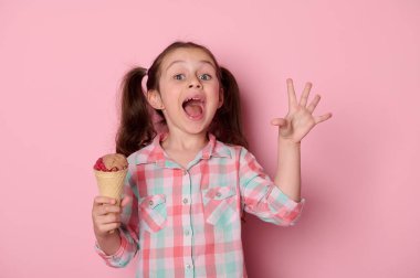 Ekose gömlekli mutlu kız çocuğu, elinde dondurmalı waffle külahı, parmaklarıyla lezzetli jest işareti yapıyor, tatlı yaz tatlısının tadını çıkarıyor, pembe arka planda izole edilmiş kameraya bakıyor,