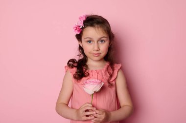 Güzel bir kız çocuğu, zarif pembe elbiseli sevimli bir kız çocuğu, elinde güzel bir gül çiçeği, kopyalanmış pembe arka planda gülümsüyor. Çocuklar işte. Çocukluk. Modaya uygun çocuklar..