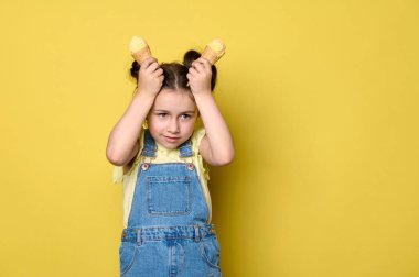 Sarı tişörtlü ve mavi kot pantolonlu sevimli yaramaz küçük kız elinde lezzetli dondurmadan yapılmış waffle konileri, surat ifadeleri, somurtmalar, eğlenmeler, sarı arka planda soyutlanmalar.