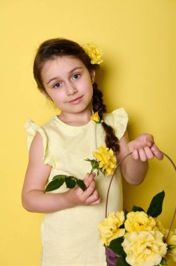 Küçük güzel bir kızın dikey portresi. Elinde mor bir hasır sepet, sarı gül çiçekleri var. Kameraya bakıp gülümsüyor. Anneler Günün kutlu olsun.