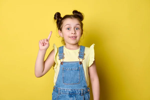 美しい驚きの小さな女の子のスタジオの肖像画は 彼女のインデックスの指を指して 孤立した黄色の背景に広告テキストのためのコピースペースを示しています 幸せな感情的な驚きの子供 女子高生 — ストック写真