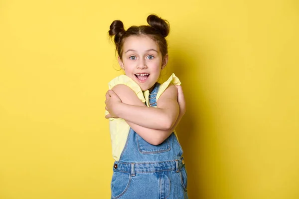 スタジオポートレートの魅力的な幸せな感情の小さな子供の女の子6歳 黄色のTシャツとブルーデニムを身に着けています 自分自身を抱きます カメラを見て笑顔 黄色の背景に肯定的な感情 — ストック写真