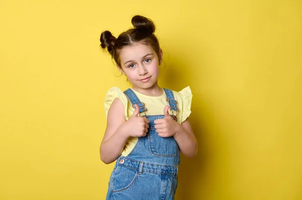 可愛いです女の子6歳 小学生ジェスチャーで親指アップ 自信を持ってカメラを見て 黄色の背景の上に立って 青いカジュアルデニム全体と黄色のTシャツを身に着けています — ストック写真