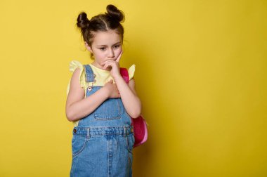 Pembe bir sırt çantası taşıyan, sarı tişörtlü, mavi kot elbiseli, süslü bir ilkokul öğrencisi kız, dudaklarına işaret parmağı koyuyor, rüya gibi bir şekilde kenara bakıyor, sarı arka plan.