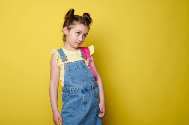 Sevimli küçük bir çocuk, birinci sınıf öğrencisi, mavi kot pantolon ve sarı tişört giymiş, pembe bir sırt çantası taşıyor, metin için bir kenara bakıyor, bir stüdyo arka planında şaşkınlık içinde