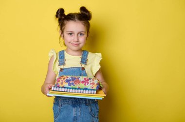 Kendine güveni olan, komik saç stili olan, okul malzemelerini elinde tutan, kameraya bakıp gülümseyen, mutlu olumlu duygularını ifade eden, yeni okul yılına başlamaya hazır, izole edilmiş sarı arka planı olan zeki kız.