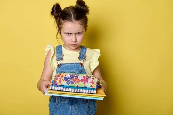 小学有感情的学生 穿着蓝色斜纹棉布工作服和黄色T恤的小女孩 满脸怒容地看着相机 拿着铅笔盒和课本 背景是黄色的 — 图库照片