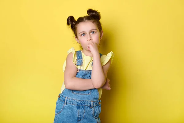 stock image Adorable little child girl standing with her arms folded, thoughtfully thinking about something, looking pensive, isolated over yellow color background. Smart kids. Primary school student. Schoolgirl