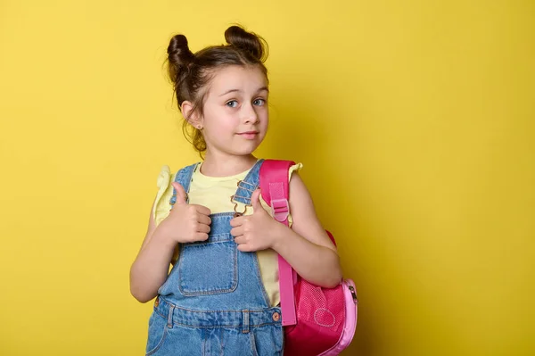 迷人的小女孩 背着粉色背包 穿着时髦的斜纹棉布工作服和黄色的T恤衫 竖起大拇指 看着相机 表达着幸福 在一年级开学时高兴极了 — 图库照片
