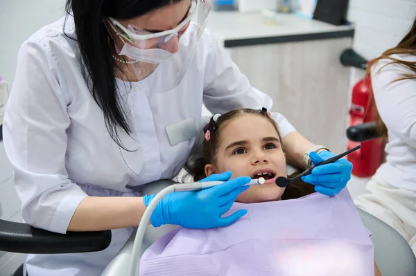 Female dentist doctor in medical protective eyewear, holding dental tools, performing dental treatment and cleaning to a little child girl in dentists chair, curing caries in dentistry clinic