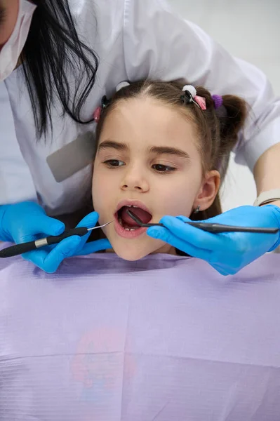 医師の歯科医によって検討されている歯科医の椅子のクローズアップ肖像画の小さな子供の女の子 歯科衛生士は歯科用具を持ち 歯の治療を行い 小児歯科診療所での治療を行う — ストック写真