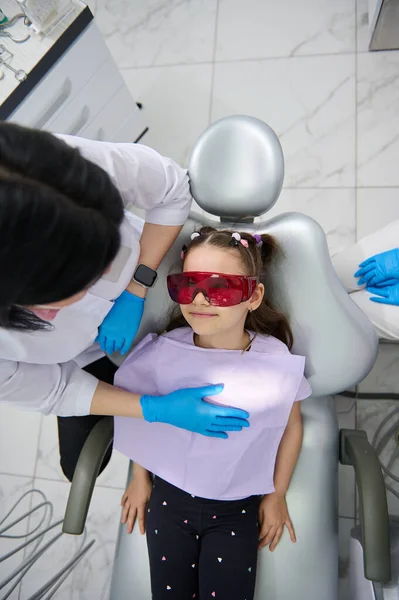 stock image Top view experienced pleasant female dentist doctor, dental hygienist talks to a little patient, adorable little child girl sitting in dentists chair during appointment in pediatric dentistry clinic