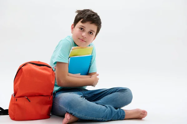 英俊的少年男孩 小学生 手里拿着课本 坐在橙色的背包旁 面带微笑 自信地看着相机 白色的背景与复制的广告空间隔离在一起 — 图库照片