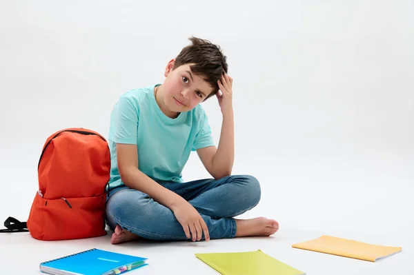 彼の頭を引っ掻いて カメラを見て 孤立した白い背景に教科書やオレンジのバックパックを配置する横に座っている 教育理念 — ストック写真