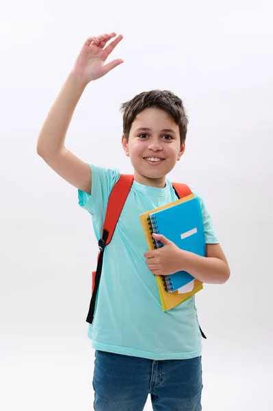 感情的な陽気な幸せな子供 スマート学童挨拶手を振って カメラを見て笑みを浮かべて オレンジ色のバックパックと学校用品を運ぶ 白い背景に孤立した 勉強だ — ストック写真