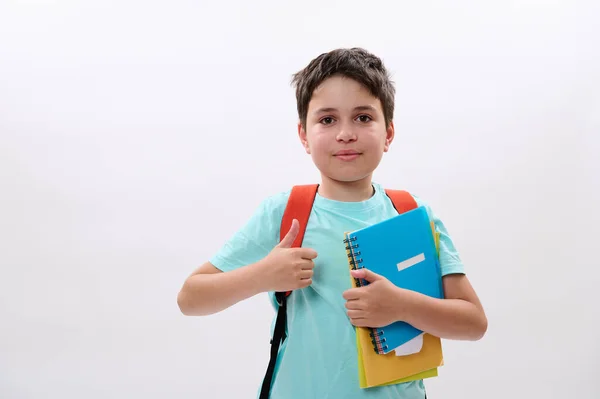 白人のスマートスクールの子供は 青いTシャツでハンサムな十代の少年は 学校用品やオレンジのバックパックを運ぶ 笑顔カメラを見て 親指を 承認サインを示しています 隔離された白い背景 — ストック写真