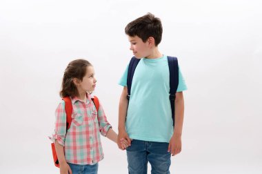 Mutlu okul çocukları, erkek ve kız kardeşler el ele tutuşuyorlar, okula gidiyorlar, gülümsüyorlar, birbirlerine bakıyorlar, beyaz arka planda soyutlanmışlar..