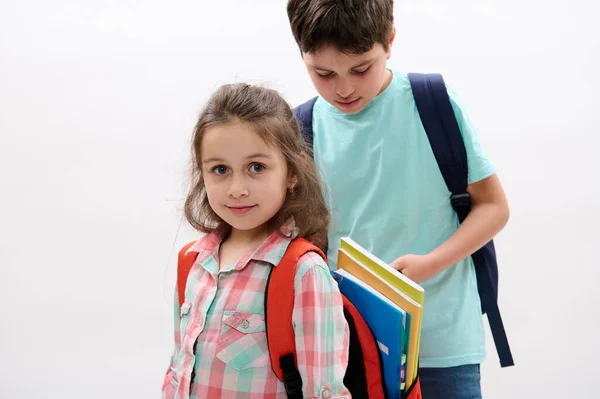快乐的小女孩 一年级 穿着格子呢休闲衫的小学生 微笑着看着相机 站在她哥哥旁边的白色背景上 把练习本放进她的背包里 — 图库照片