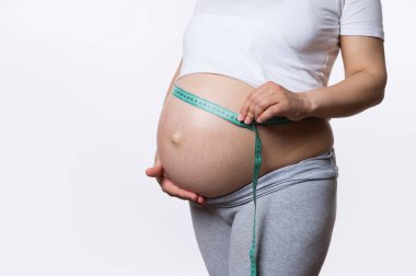 Hamile bir kadının göbeğini bir bant ile ölçerek fetüs gelişimini takip etmesi, beyaz stüdyo arka planında izole edilmiş. Sağlıklı hamilelik. Kadın doğum ve jinekoloji konsepti