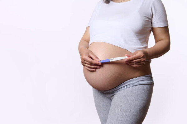 Close-up gravid woman, expectant mother holding a pregnancy inkjet test kit above her pregnant belly, isolated on white background. Fertility. Womens health. Child expectation. Pregnancy. Maternity