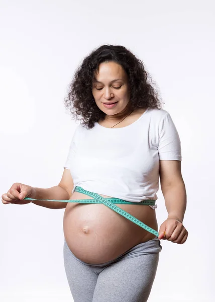 一个卷曲头发的成年孕妇 准妈妈的广告演播室肖像 监测她胎儿在子宫中的发育 用带子测量她的大肚子 用白色背景隔开 — 图库照片